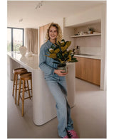Eine lächelnde Person in Jeanskleidung steht in einer modernen Küche und hält eine künstliche Magnolie in einem 46-cm-Topf, eine pflegeleichte Pflanze. Die Küche eignet sich perfekt für die Inneneinrichtung und verfügt über Holzschränke, eine weiße Kücheninsel und zwei Barhocker.