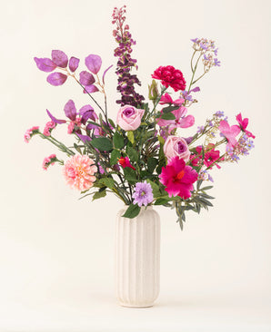 Eine weiße strukturierte Vase enthält den künstlichen Seidenblumenstrauß Purple Dawn in Rosa-, Lila- und Lavendeltönen mit grünem Blattwerk vor einem hellen, neutralen Hintergrund.