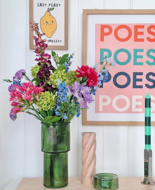Eine grüne Glasvase hält einen künstlichen Seidenblumenstrauß Blossom Breeze auf dem Tisch. Dahinter hängen gerahmte Bilder – eines mit einer Zitrone und „EASY PEASY“, ein weiteres mit „POESY“ – für zusätzlichen Charme. In der Nähe stehen eine grüne Kerze und eine rosa Spiralkerze von Blossom Breeze.
