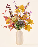 Der Strauß „Wilder Amethyst“ aus künstlichen Seidenblumen mit Herbstblättern, violetten und weißen Blüten und grünen Hortensien steht in einer weißen Keramikvase vor einem schlichten, hellen Hintergrund.