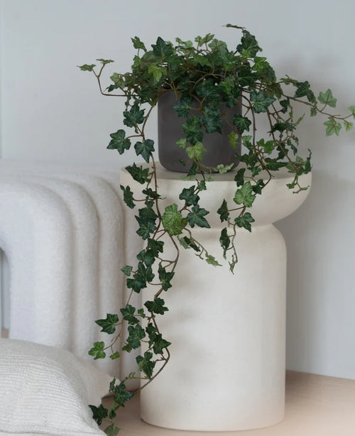 Ein künstlicher Efeu Deluxe Green, ein 90 cm großer hängender Efeu in einem grauen Topf, steht auf einem modernen, weißen, skulpturalen Sockel neben einer gefalteten weißen Decke und einem weißen Strukturkissen.