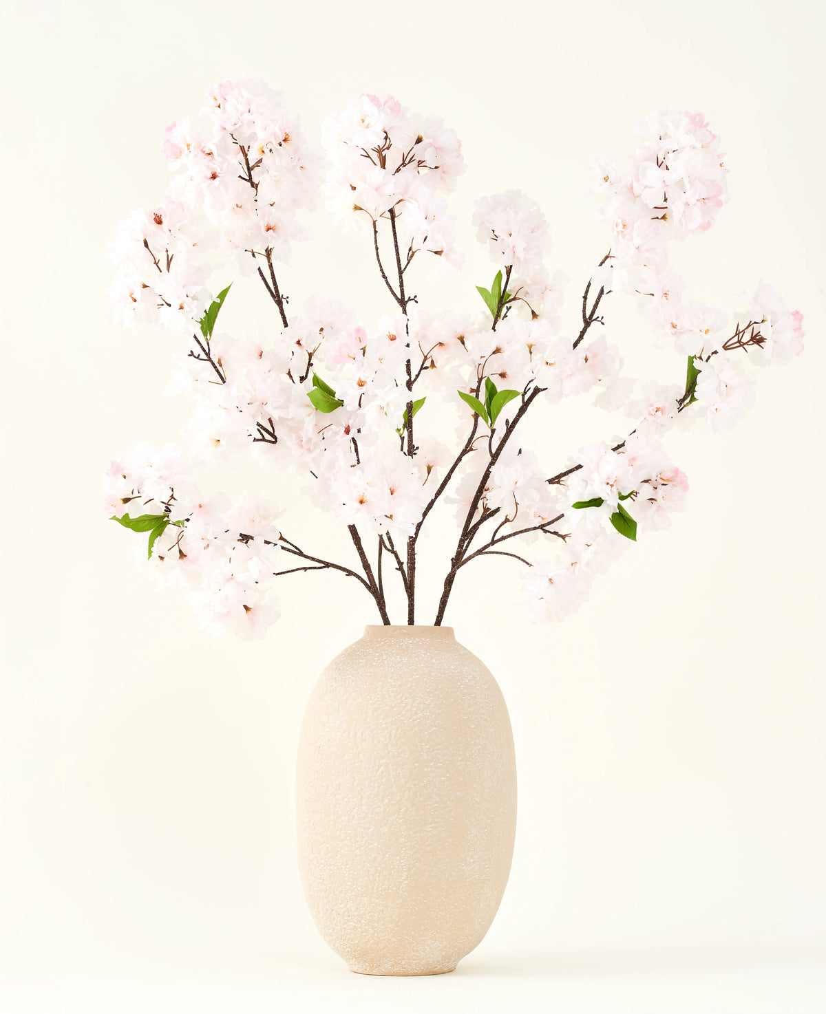 Der Seiden-Kunstblumenstrauß Cherry Blossom Blush 119 cm zeigt zarte, hellrosa Kirschblüten mit grünen Blättern in einer beigen Vase, ideal als künstliche Blumendekoration vor hellem Hintergrund.