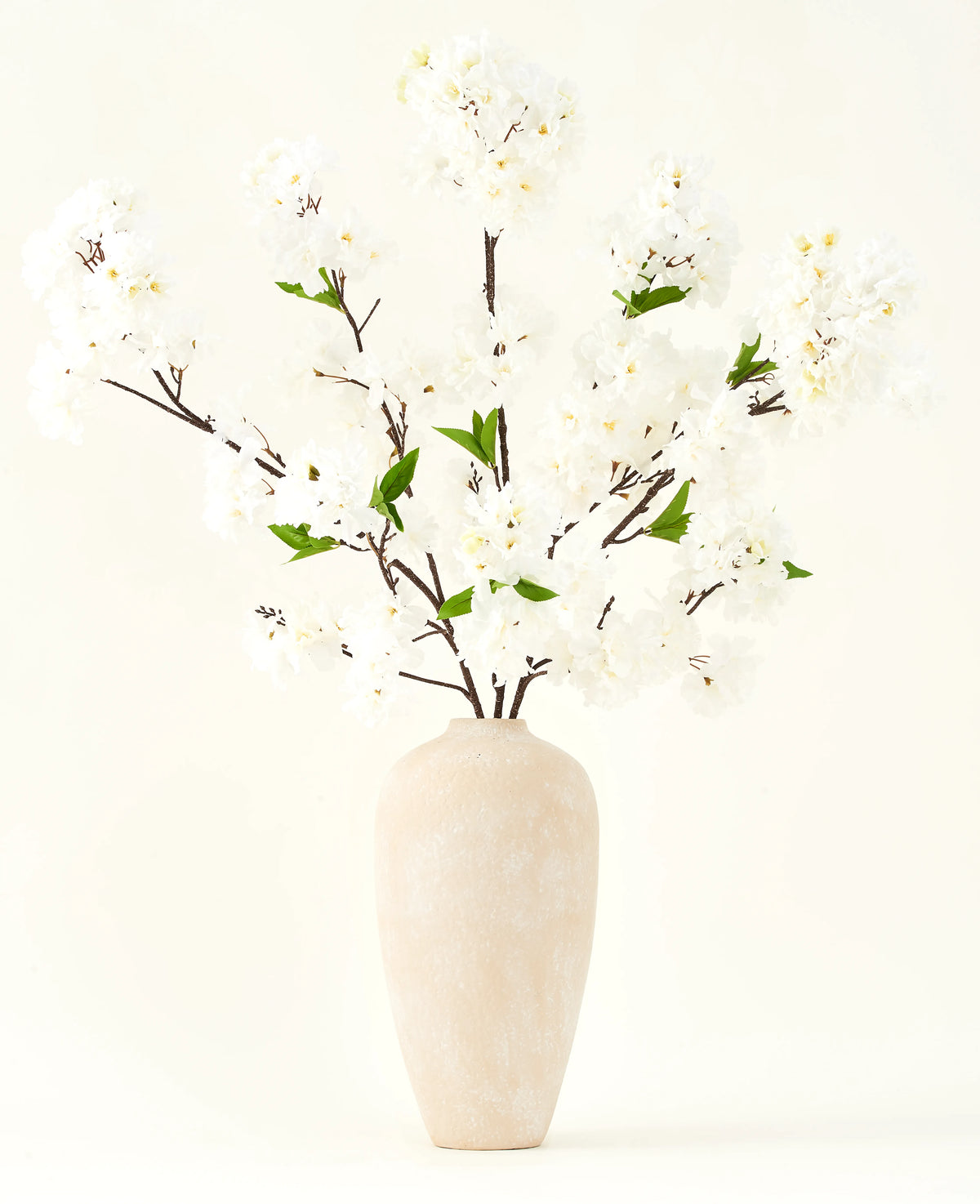 Der künstliche Seidenblumenstrauß Kirschblütencreme 119 cm besteht aus eleganten braunen Zweigen mit zartweißen Kunstblüten und grünen Blättern, die in einer schlichten beigen Vase vor einem schlichten hellen Hintergrund präsentiert werden.