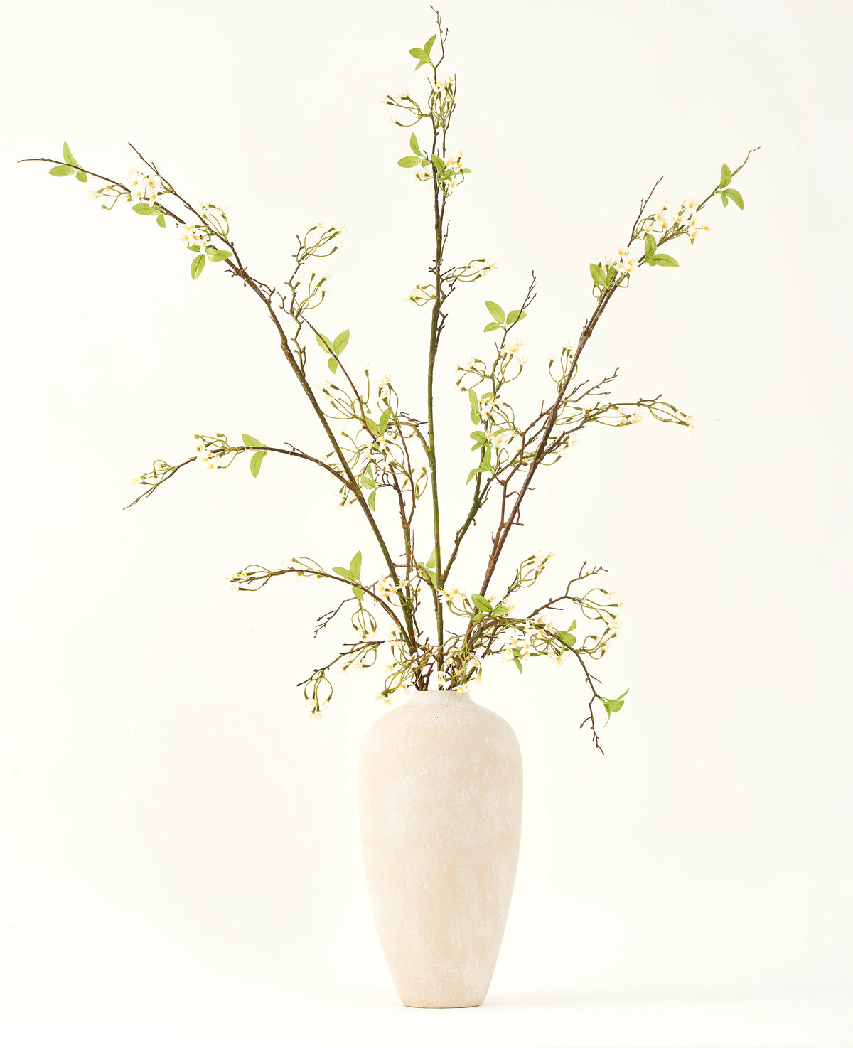 In einer hohen, cremefarbenen Keramikvase befindet sich der künstliche Seidenblumenstrauß Verbena Creme mit langen Zweigen, kleinen grünen Blättern und zarten weißen Blüten, wunderschön arrangiert vor einem schlichten hellen Hintergrund.