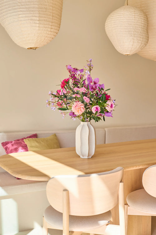 Auf einem Esstisch aus hellem Holz steht eine weiße Vase mit dem künstlichen Seidenblumenstrauß Purple Dawn, der vor leuchtenden Rosa- und Lilatönen strotzt. Dahinter vervollständigen eine gepolsterte Bank mit Kissen und zwei hängende Papierlaternen das Bild und verleihen dem Raum die ruhige, von Purple Dawn inspirierte Atmosphäre.