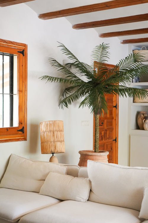 Ein gemütliches Wohnzimmer mit einem weißen Sofa, einer Rattan-Tischlampe, der Kunstpalme Phoenix De Luxe 210 cm im Topf, Holzbalken an der Decke, einem Fenster mit Holzrahmen und einer Holztür im Hintergrund.