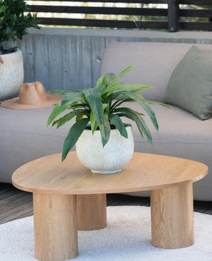 Eine 60 cm große Kunstpflanze Bird's Deluxe steht auf einem runden Couchtisch aus Holz vor einem beigen Sofa, dekoriert mit einem grünen Kissen und einem braunen Hut, der über der Armlehne hängt.