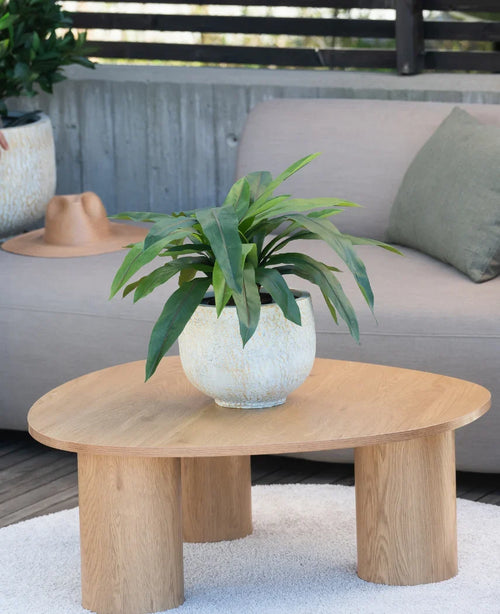 Eine 60 cm große Kunstpflanze Bird's Deluxe steht auf einem runden Couchtisch aus Holz vor einem beigen Sofa, dekoriert mit einem grünen Kissen und einem braunen Hut, der über der Armlehne hängt.
