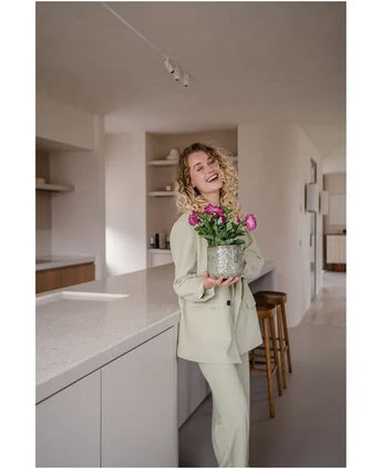 Eine lächelnde Frau mit lockigem Haar und hellem Anzug steht in einer modernen, minimalistischen Küche und hält eine künstliche Petunie UV 25 cm dunkelrosa mit leuchtend rosa Blüten in der Hand.