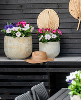 Zwei große Keramiktöpfe mit einer künstlichen Petunie UV 25 cm dunkelrosa, mit leuchtend rosa Blüten, stehen auf einem Tisch vor einer dunklen Holzwand. Auf dem Tisch steht ein Strohhut und über den Blumen hängen getrocknete Palmblätter an der Wand.