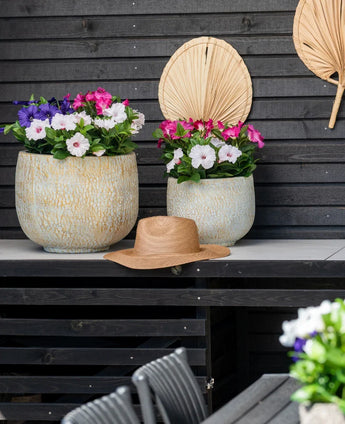 Zwei große Keramiktöpfe mit einer künstlichen Petunie UV 25 cm dunkelrosa, mit leuchtend rosa Blüten, stehen auf einem Tisch vor einer dunklen Holzwand. Auf dem Tisch steht ein Strohhut und über den Blumen hängen getrocknete Palmblätter an der Wand.