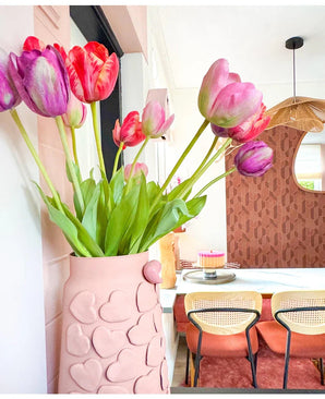 Een vaas met roze hartmotieven houdt Kunstbloem Franse Tulp 60 cm paars op een eettafel met stoelen met geweven rugleuning. Op de achtergrond dragen een ronde spiegel en een muur met bruin patroon bij aan het gezellige, moderne interieur.