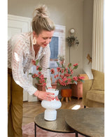 Eine Frau mit blonden Haaren in einem Dutt arrangiert die Kunsttak Berries 64 cm rosa in einer weißen Vase auf einem runden Tisch in einem gemütlichen Wohnzimmer mit Erdtönen und moderner Einrichtung.