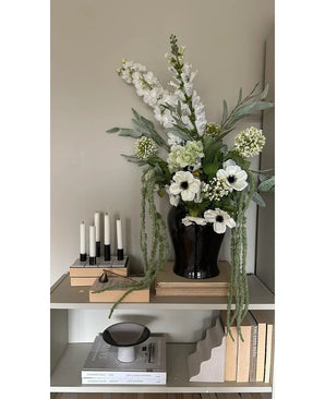 Auf einem Regal an einer hellen Wand steht eine schwarze Vase mit der Kunstblume Amaranthus 124 cm grün, daneben fünf weiße Kerzen, beige Bücher und Dekogegenstände in neutralen Tönen.