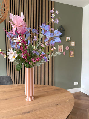 Eine hohe rosa-weiß gestreifte Vase mit dem künstlichen Seidenblumenstrauß Amethyst Romance steht auf einem runden Holztisch in einem modernen Raum mit grünen Wänden, Holzvertäfelung und kleinen gerahmten Kunstwerken.