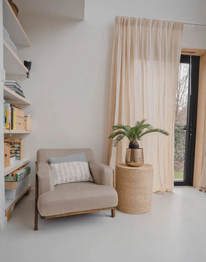Eine gemütliche Leseecke mit einem beigen Sessel, einem runden Beistelltisch aus Korbgeflecht mit der künstlichen Baby-Cycas-Palme 60 cm darauf, Bücherregalen und Gardinen, die über einer Glastür hängen, die zum Garten führt.