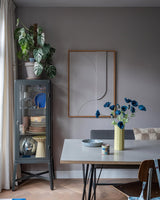 Der moderne Essbereich verfügt über eine graue Glasvitrine, in der Geschirr und Pflanzen ausgestellt sind, minimalistische Wanddekorationen und einen Tisch, auf dem die Kunstblume Mohn 73 cm dunkelblau in einer gelben Vase steht. Abgerundet wird der Raum durch eine eklektische Mischung aus Stühlen, alle vor dem Hintergrund grauer Wände und eines Holzbodens.