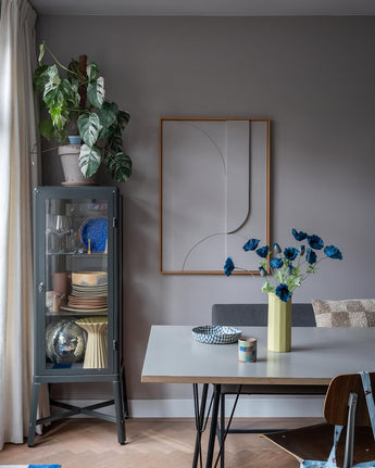 Der moderne Essbereich verfügt über eine graue Glasvitrine, in der Geschirr und Pflanzen ausgestellt sind, minimalistische Wanddekorationen und einen Tisch, auf dem die Kunstblume Mohn 73 cm dunkelblau in einer gelben Vase steht. Abgerundet wird der Raum durch eine eklektische Mischung aus Stühlen, alle vor dem Hintergrund grauer Wände und eines Holzbodens.