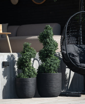 Zwei künstliche Buchsbaumspiralen im römischen UV-Stil (90 cm) mit Spiralblättern stehen auf einer Betonterrasse neben einem schwarzen Hängesessel und einem modernen Außensofa im Hintergrund.