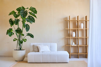 Minimalistische woonkamer met een Kunstplant Monstera 240 cm in een pot, een moderne witte bank met kussen en een houten open kast met witte vazen tegen een beige muur. Natuurlijke lichtinval van rechts.