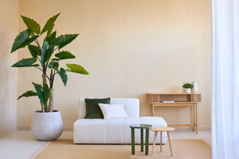 Een minimalistische woonkamer met de Kunstplant Calocasia 210 cm, een bank met witte textuur en groene en witte kussens, twee kleine bijzettafeltjes, een beige vloerkleed en een houten consoletafel tegen een lichtbeige muur.