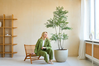 Eine Frau in einem grünen Anzug lächelt, während sie auf einem Holzstuhl neben der künstlichen Ruscus-Pflanze (180 cm) in einem hellen, modernen Raum mit beigen Wänden, großen Fenstern und minimalistischem Dekor sitzt.