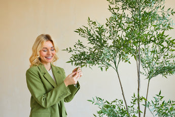 Eine lächelnde Frau in einem grünen Blazer steht drinnen und berührt sanft die Blätter einer 150 cm großen künstlichen Ruscus-Pflanze mit dünnen Zweigen und üppig grünem Laub, die sich vor einem schlichten, hellen Hintergrund abhebt.
