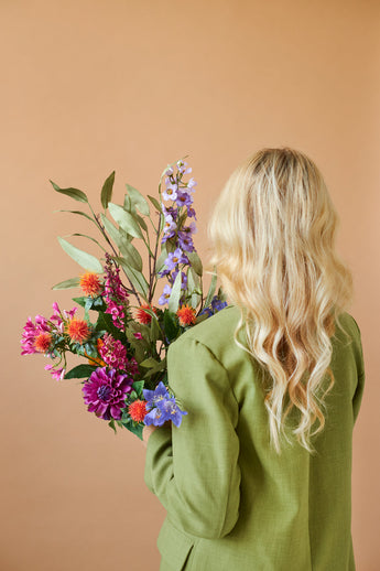Von hinten ist eine Person mit langen, welligen blonden Haaren und einer grünen Jacke zu sehen, die den farbenfrohen Kunstblumenstrauß aus Svenski-Seide vor einem schlichten beigen Hintergrund hält.