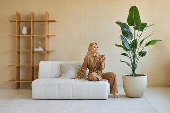 Eine Frau in braunem Outfit sitzt lächelnd auf einer weißen Couch und hält eine Tasse in der Hand. Neben ihr steht die Kunstpflanze Strelitzia 180 cm Real Touch, die mit minimalistischer Ausstattung üppiges Grün in das moderne, beige getönte Wohnzimmer bringt.
