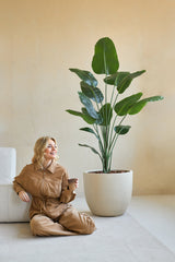 Eine Frau in einem braunen Overall sitzt lächelnd auf dem Boden, neben sich eine Kaffeetasse, neben der Kunstpflanze Strelitzia 180 cm echte Hingucker in einem minimalistischen Raum mit beigen Wänden.