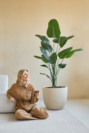 Eine Frau in einem braunen Overall sitzt lächelnd auf dem Boden, neben sich eine Kaffeetasse, neben der Kunstpflanze Strelitzia 180 cm echte Hingucker in einem minimalistischen Raum mit beigen Wänden.