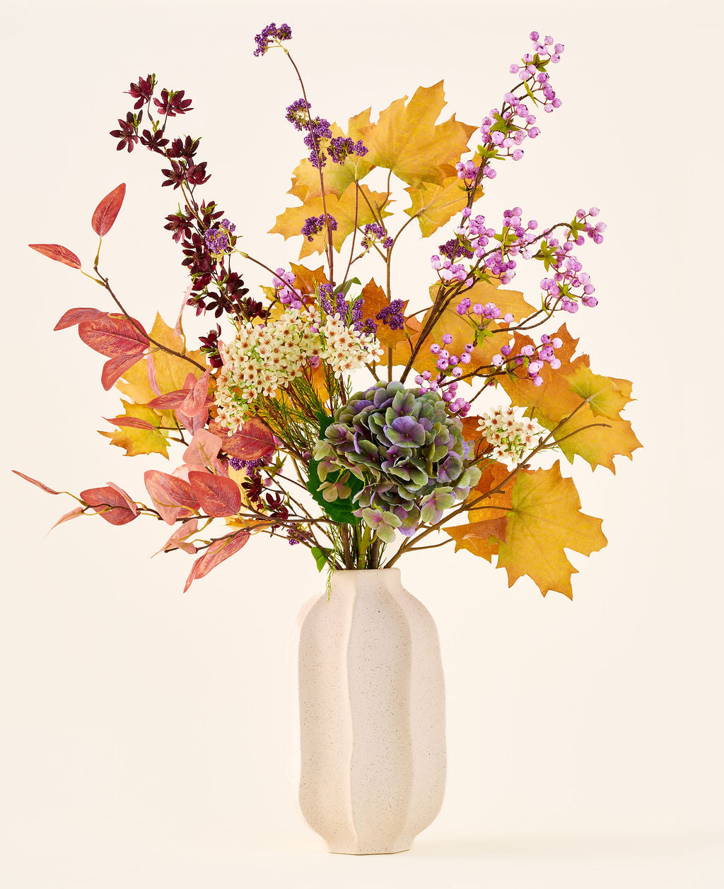 Der Strauß „Wilder Amethyst“ aus künstlichen Seidenblumen mit Herbstblättern, violetten und weißen Blüten und grünen Hortensien steht in einer weißen Keramikvase vor einem schlichten, hellen Hintergrund.