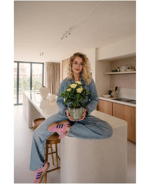 Een vrouw met krullend blond haar, gekleed in spijkerkleding en roze sneakers, zit in kleermakerszit op een keukeneiland in een moderne, zonovergoten keuken, met de Kunstplant Gardenia UV 39 cm in haar handen.