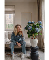Een vrouw met krullend blond haar, gekleed in spijkerkleding, zit op een beige bank en staart naar een Kunstplant Hortensia UV 85 cm blauw die op een zwarte standaard staat. De kamer straalt rust uit, is zacht verlicht en heeft op de achtergrond een raam.
