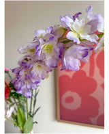 Nahaufnahme der künstlichen Blume Delphinium 105 cm lila, arrangiert in einer Vase, mit einem rosa und roten Blumenkunstwerk an der Wand im Hintergrund.