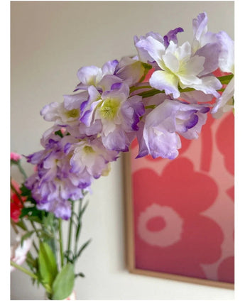 Nahaufnahme der künstlichen Blume Delphinium 105 cm lila, arrangiert in einer Vase, mit einem rosa und roten Blumenkunstwerk an der Wand im Hintergrund.