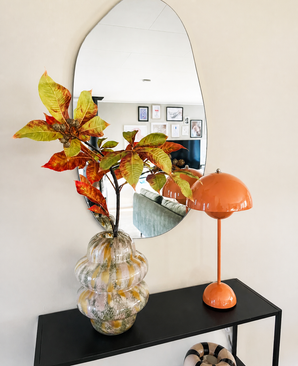 In een golvende spiegel boven een zwarte consoletafel staan een glazen vaas met Kunsttak Kastanje 91 cm rood/groen en een oranje paddestoellamp. De spiegel weerspiegelt een woonkamer met ingelijste kunst aan de muur.