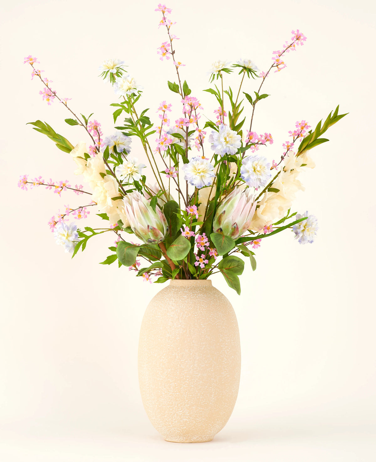 In einer Vase mit beiger Struktur befindet sich der Seiden-Kunstblumenstrauß Lots of Love XL, ein handgebundener Strauß aus rosa, weißen und blauen Seidenblumen mit grünen Blättern und langen Stielen vor hellem Hintergrund.