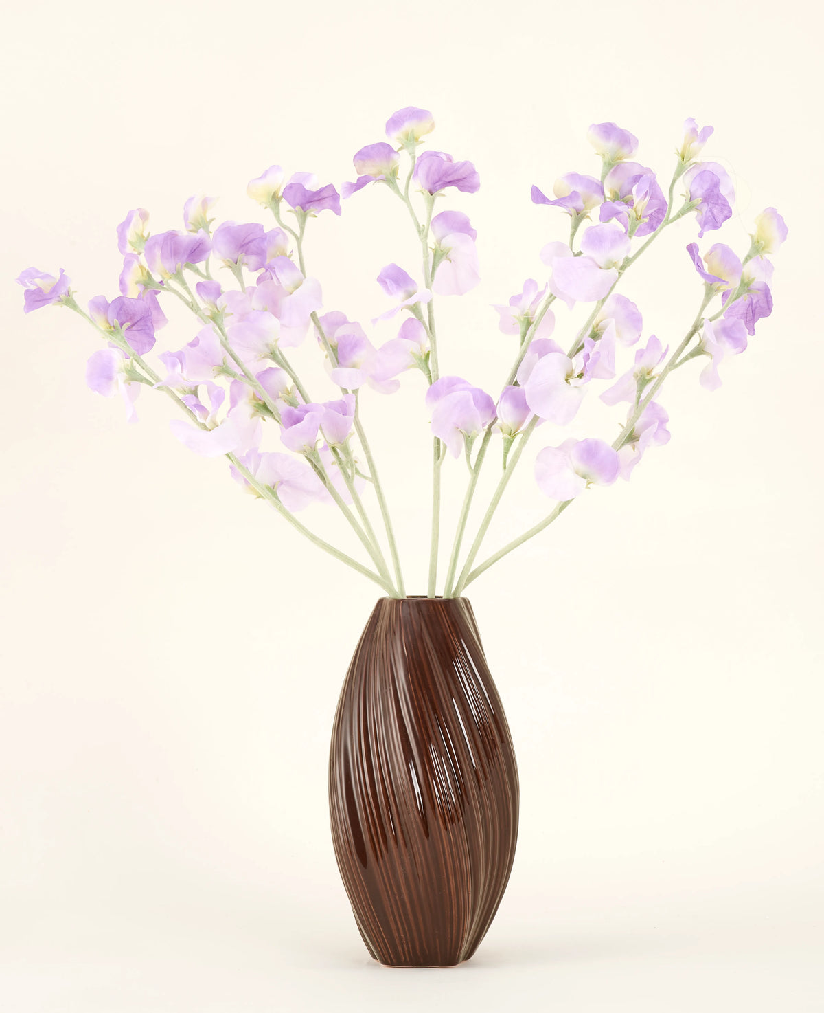 Eine strukturierte braune Vase hält den künstlichen Seidenblumenstrauß Erbsenlila-hellviolette Seidenblumen mit grünlichen Stielen vor einem schlichten, hellen Hintergrund.