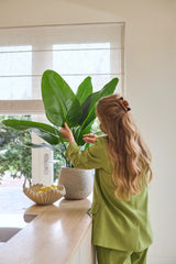 Een vrouw met lang golvend haar, gekleed in een groen pak, verzorgt een Kunstplant Strelitzia Real Touch Deluxe van 90 cm op een toonbank naast een schaal met citroenen in een lichte kamer met een raam.
