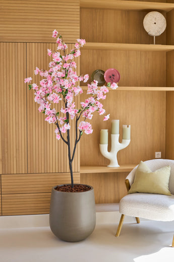 Een Kunst Kersenbloesem 180 cm roze, een potboom met roze bloesems, staat naast een witte stoel met kussens in een moderne kamer met houten planken en decoratieve voorwerpen.