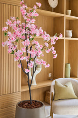 De Kunst Kersenbloesem 180 cm roze, met zijn delicate roze bloesems, staat in een moderne woonkamer met houten planken, keramiek, een beige fauteuil met een groen kussen en een minimalistische inrichting.