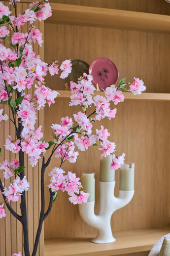Een Kunst Kersenbloesem van 180 cm in roze staat voor houten planken waarop een roze bord, een groene schaal en een witte kandelaar met drie lichtgroene kaarsen staan uitgestald.