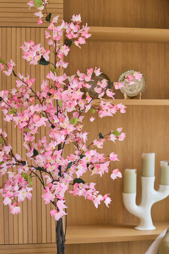 Eine künstliche Bougainvillea-Pflanze von 180 cm, rosafarben mit rosa Blüten, steht vor Holzregalen, auf denen ein dekorativer Teller und ein weißer Kerzenständer mit drei grünen Kerzen stehen.