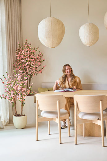 Een vrouw met lang blond haar zit aan een lichte houten tafel, een boek te lezen en glimlachend. Naast haar staat de Kunstplant Kersenbloesem 150 cm roze, waarvan de roze bloesems een levendig accent geven. Hanglampen en een minimalistische inrichting zorgen voor een lichte, gezellige sfeer.