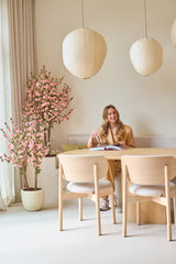 Een vrouw zit glimlachend aan een tafel met een open boek. De kamer is ingericht met lichte houten meubels, papieren lantaarns, zacht natuurlijk licht dat door hoge gordijnen naar binnen valt, en een Kunstplant Kersenbloesem 120 cm roze die een vleugje roze bloesem toevoegt.