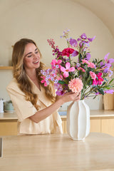 Eine lächelnde Frau mit langen blonden Haaren stellt einen bunten Strauß „Purple Dawn“ aus künstlichen Seidenblumen in einer weißen Vase auf die Theke.