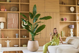 Een vrouw in een groene outfit zit op een beige bank met een drankje in haar hand, met de Kunstplant Strelitzia Nicolai Deluxe 210 cm naast haar en houten planken met decoratieve voorwerpen op de achtergrond.