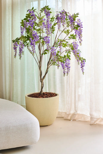 De Kunstplant Blauweregen 210 cm, met zijn overhangende trossen paarse bloemen, staat binnenshuis naast doorschijnende beige gordijnen en een lichtgekleurde bank.