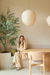 Een vrouw in een beige pak zit glimlachend aan een houten tafel in een moderne, minimalistische kamer met lichte muren, papieren hanglampen die aan het plafond hangen en een Kunstplant Eucalyptus van 180 cm die de ruimte wat groen geeft.